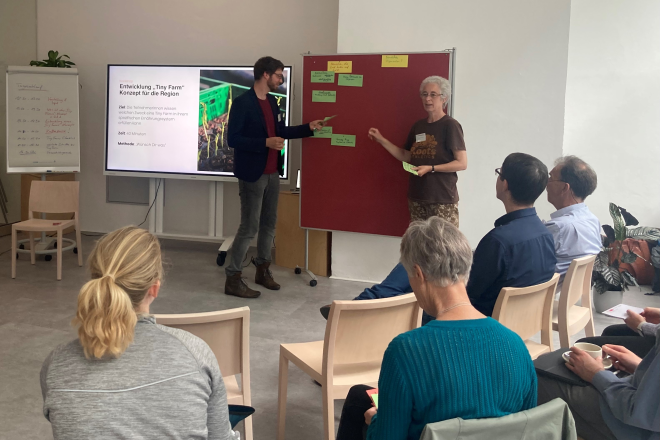 Ein Mann steht neben einer roten Pinnwand und präsentiert Informationen vor einem Publikum. Eine weitere Person, eine Frau, zeigt auf die Pinnwand, während die sitzenden Teilnehmenden aufmerksam zuschauen. Auf der Präsentationsfolie steht "Entwicklung 'Tiny Farm' Konzept für die Region."